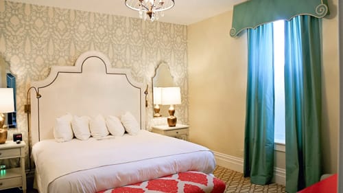 Hotel bedroom with white upholstered headboard, chandelier and teal drapes.