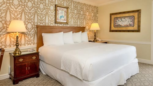 Hotel king bed with wood headboard, patterned wallpaper and framed artwork.